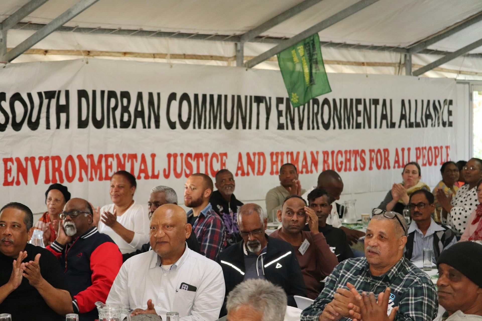 A group of people sit in a marquee with a South Durban Community Environmental Alliance banner on the wall that also says 'Environmental Justice and Human Rights for all People'