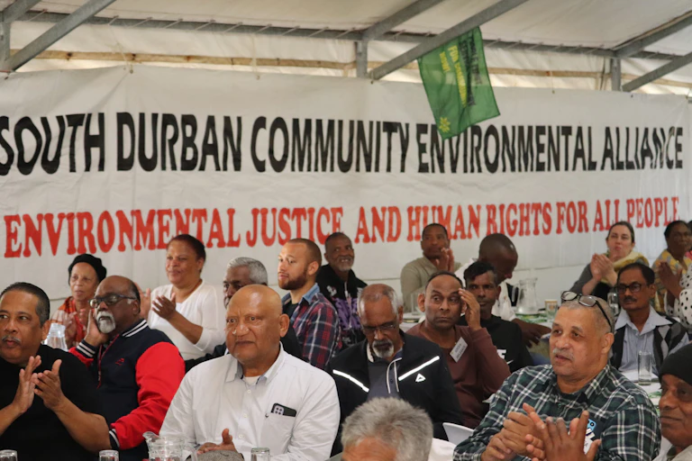A group of people sit in a marquee with a South Durban Community Environmental Alliance banner on the wall that also says 'Environmental Justice and Human Rights for all People'