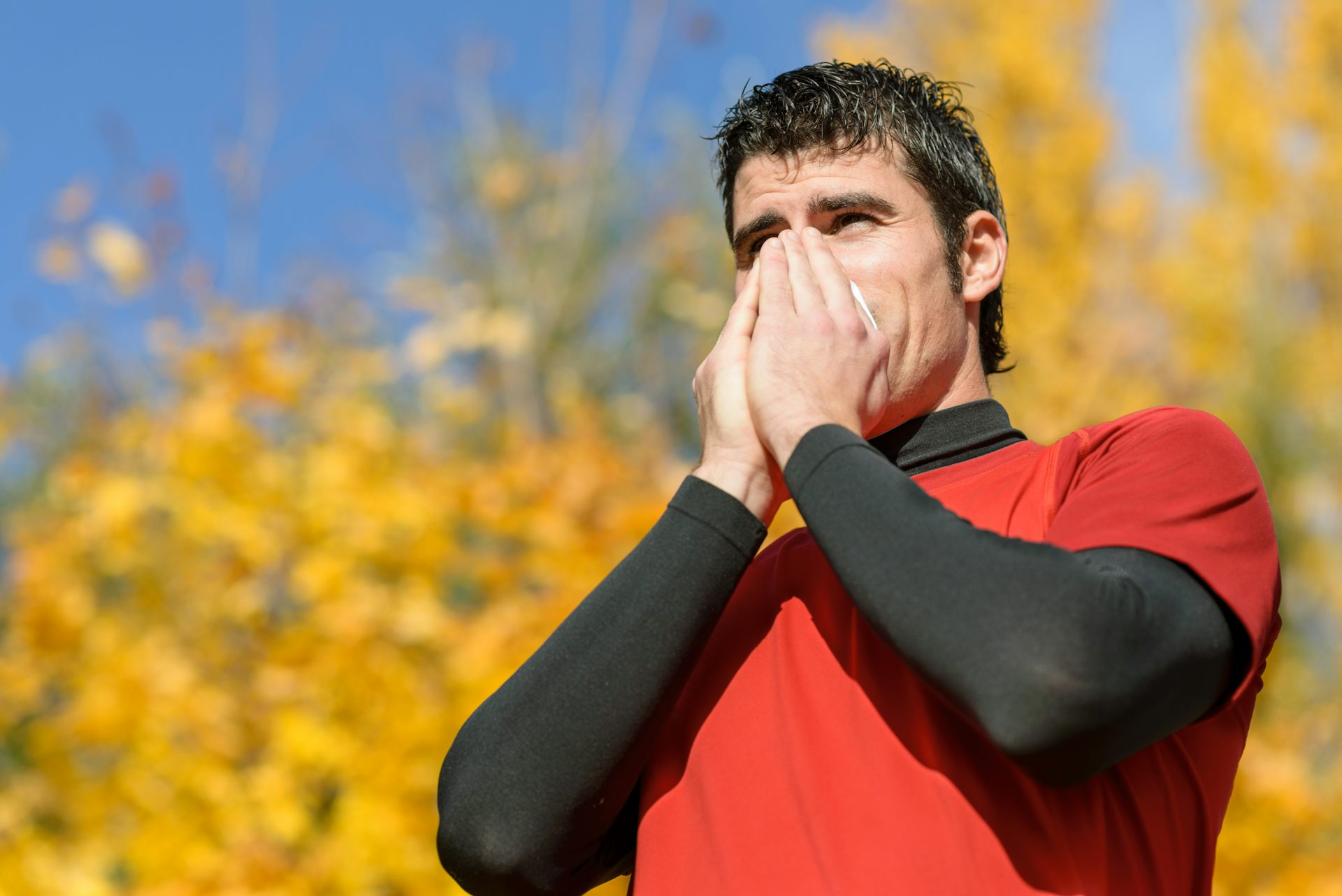 A man standing outside wearing workout clothes blows his nose into a tissue.