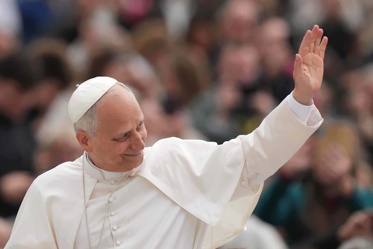A man in white robes and a white skullcap waves to a large crowd.
