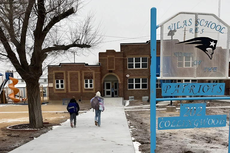 Two children walk toward a large brown brick school building.