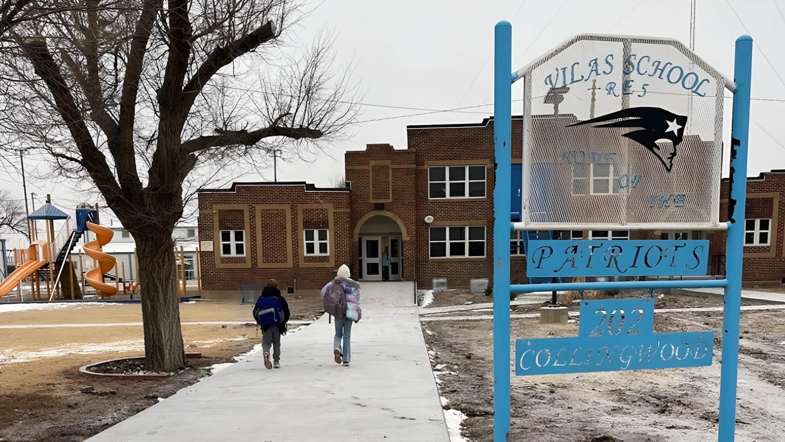 Two children walk toward a large brown brick school building.