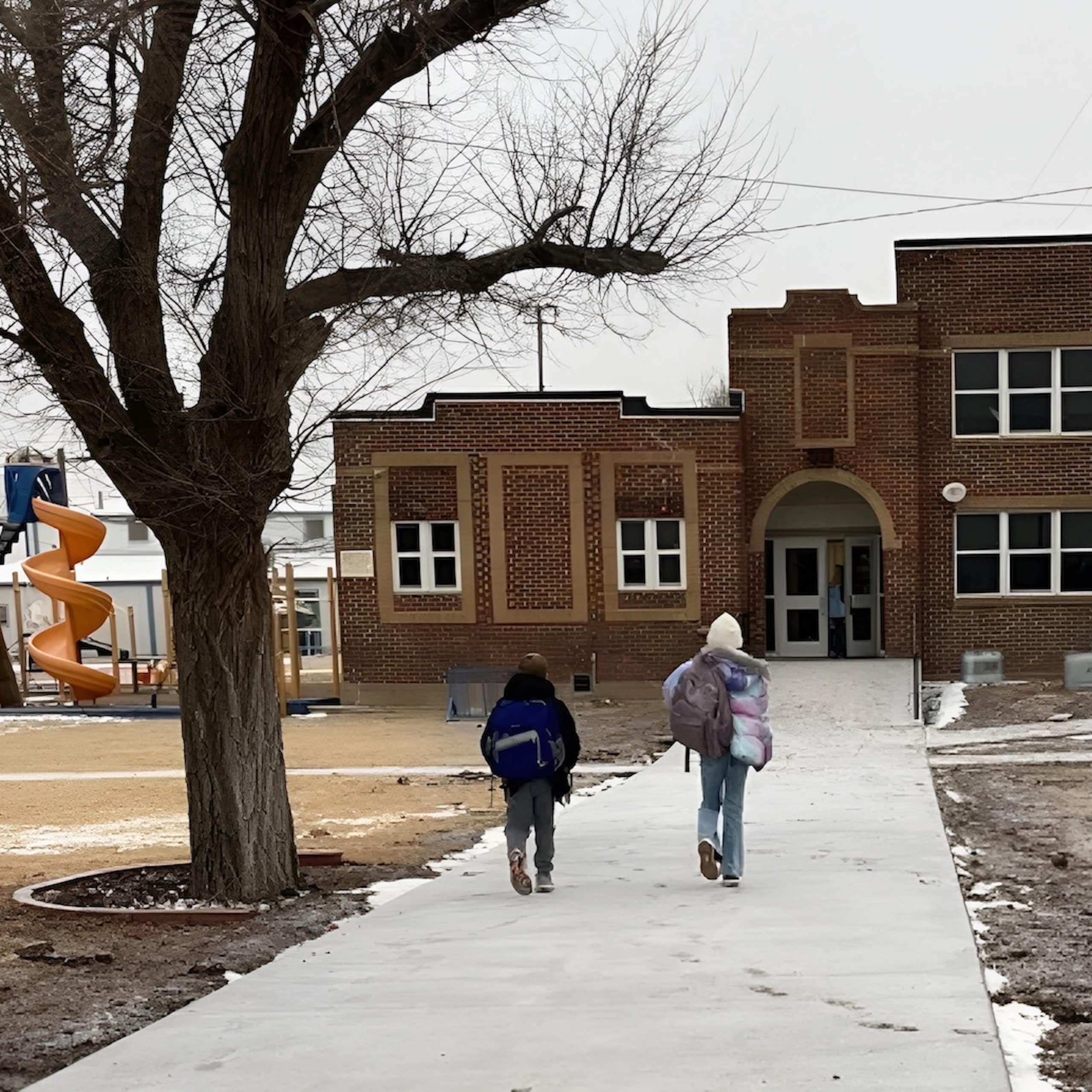 Two children walk toward a large brown brick school building.