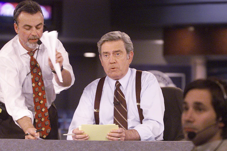A man in a shirt and tie sits at a news desk, holding cue cards, with a producer standing beside him.