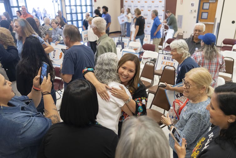 A woman hugs another woman in a room full of people.