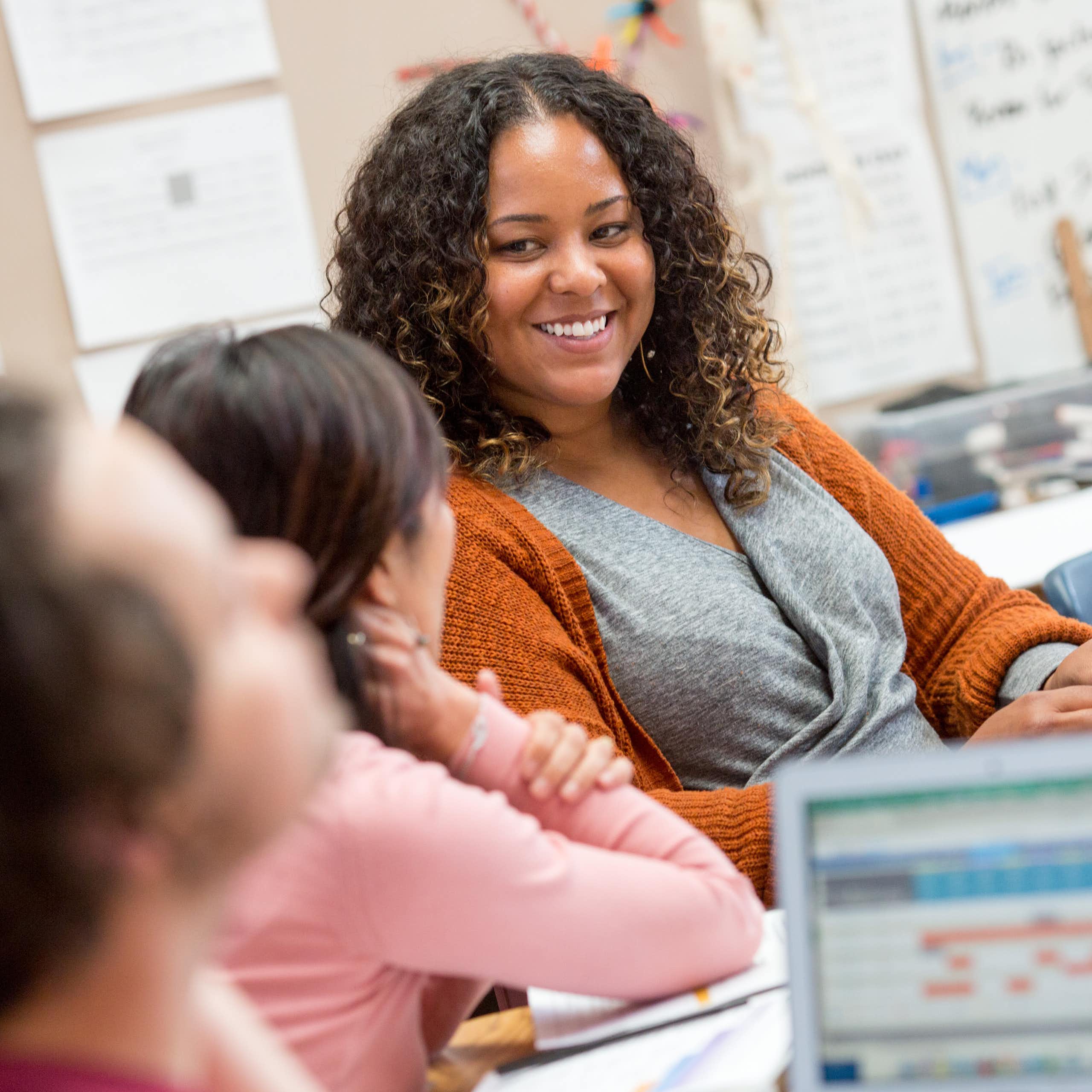(A teacher smiles at some students near a laptop on a desk)