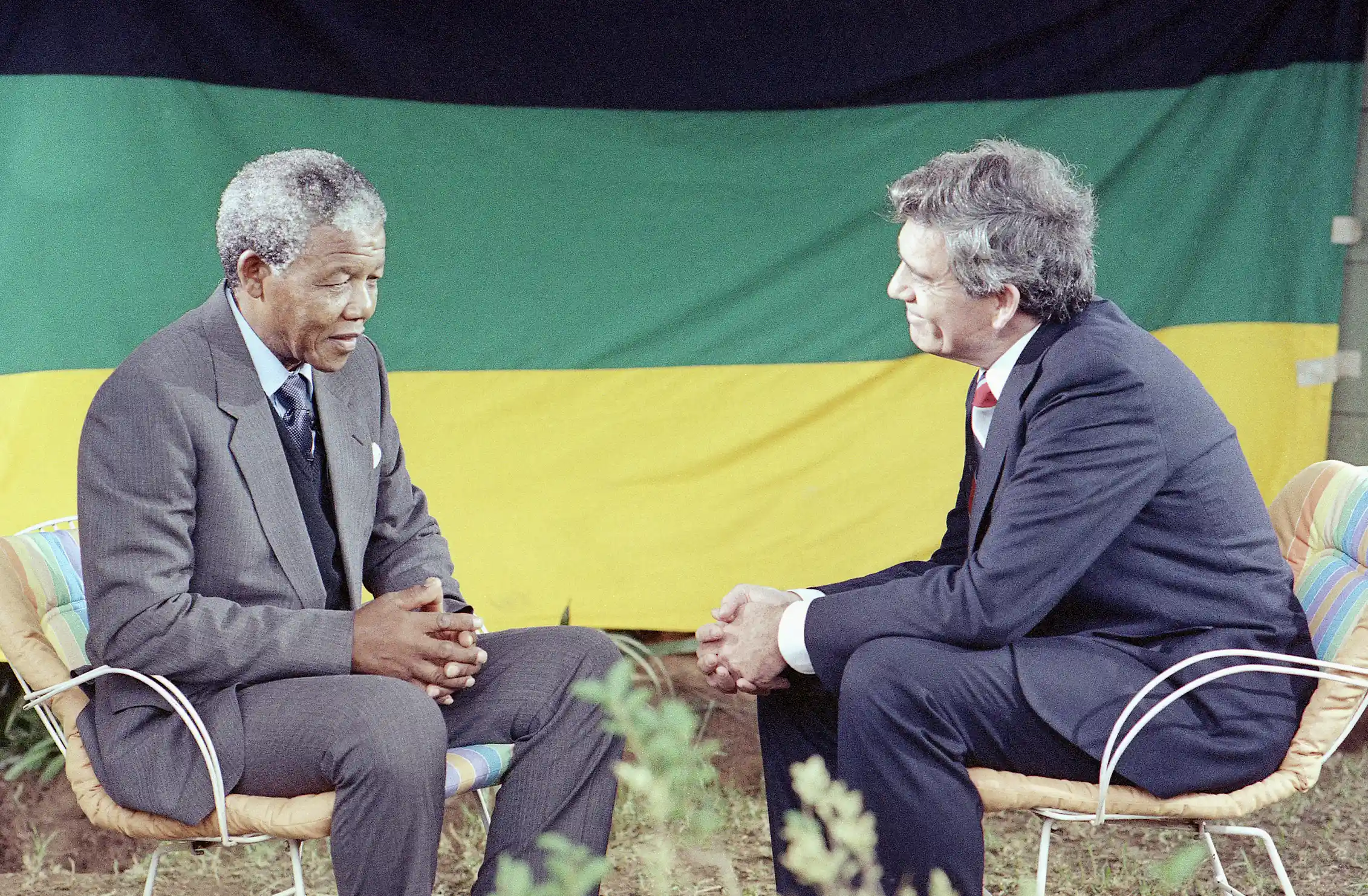 Two men sit in chairs facing each other, with the African National Congress flag behind them.