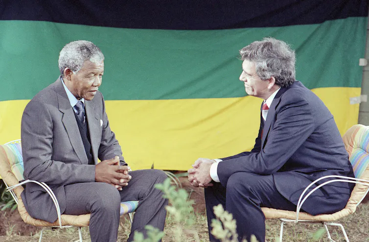Two men sit in chairs facing each other, with the African National Congress flag behind them.