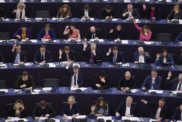 Rows of people sitting in an auditorium raise their hands on a vote