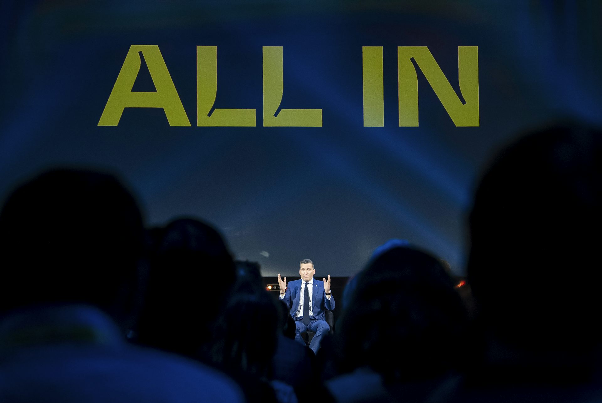 A man sits on a stage under a screen that says 'All in'