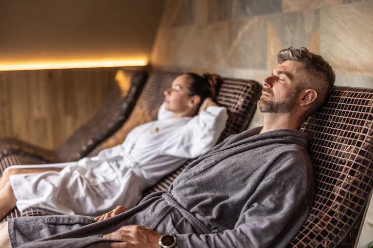 Man and woman wearing bathrobes lying on lounges, eyes shut, in a wellness spa