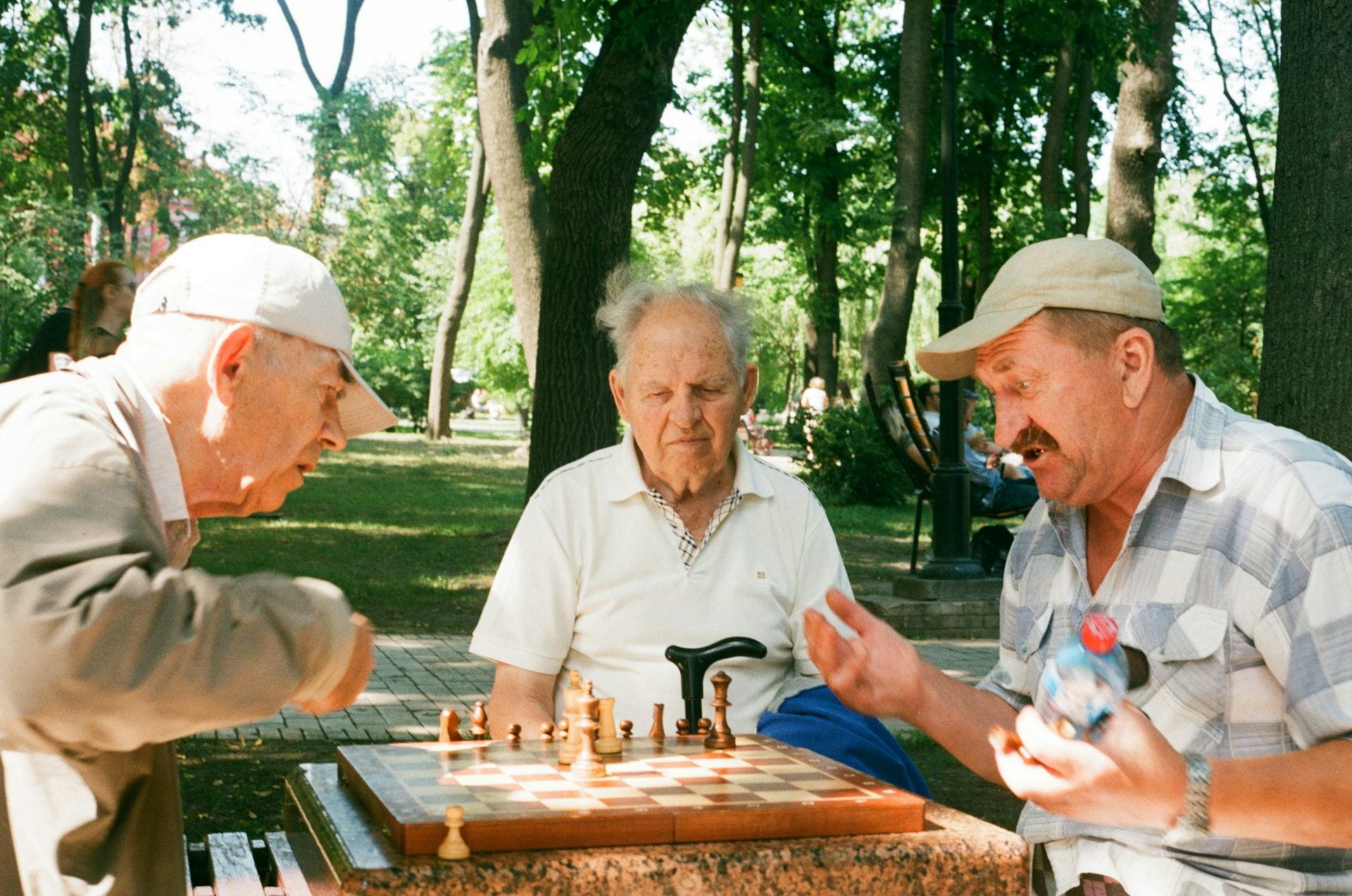 Three men with grey hair sitting at an outdoor chess table