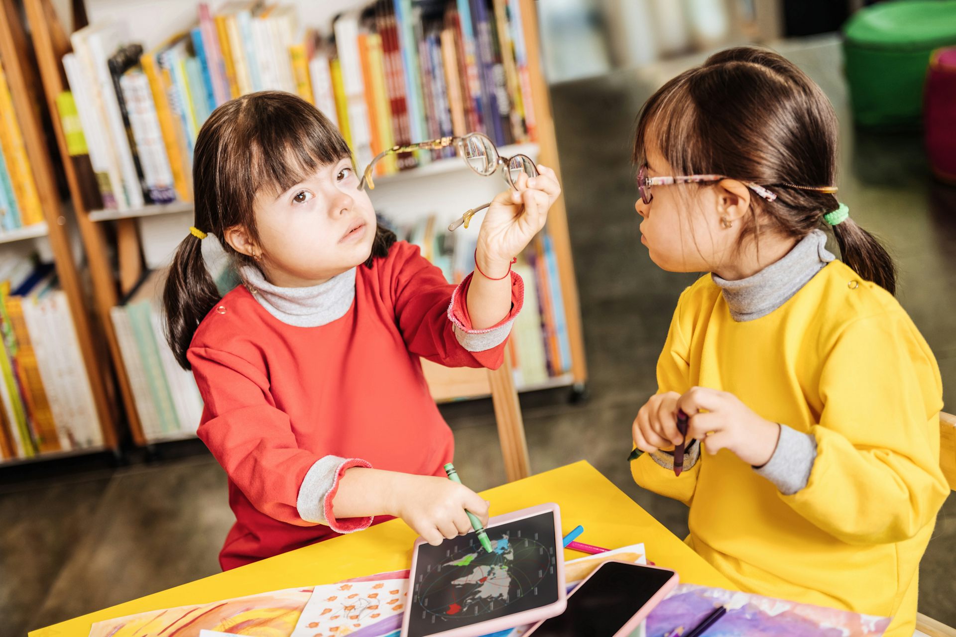 Two children sitting at a table coloring, one holding glasses aloft