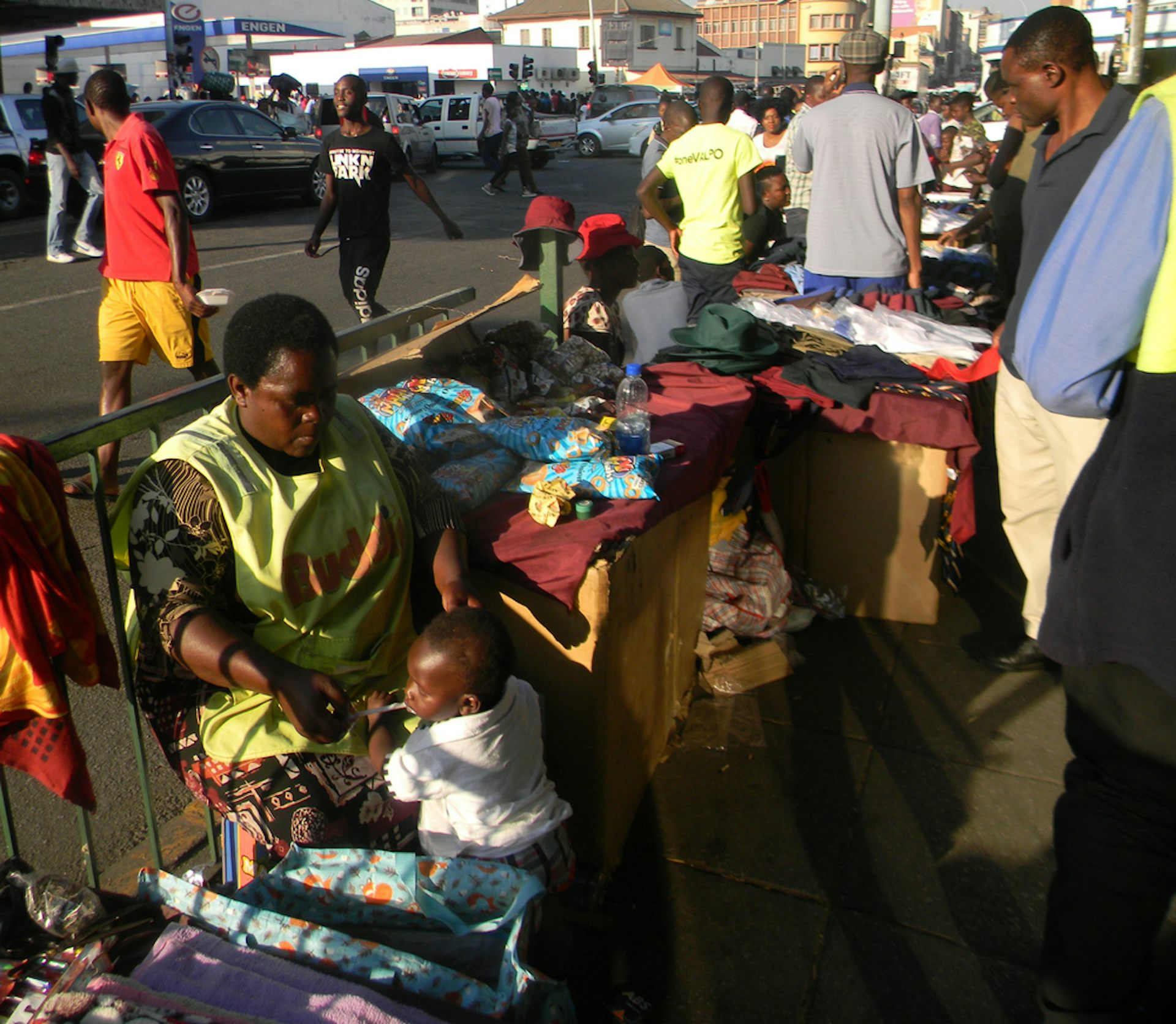 Street scene with pedestrians and a woman seated with a display of clothing
