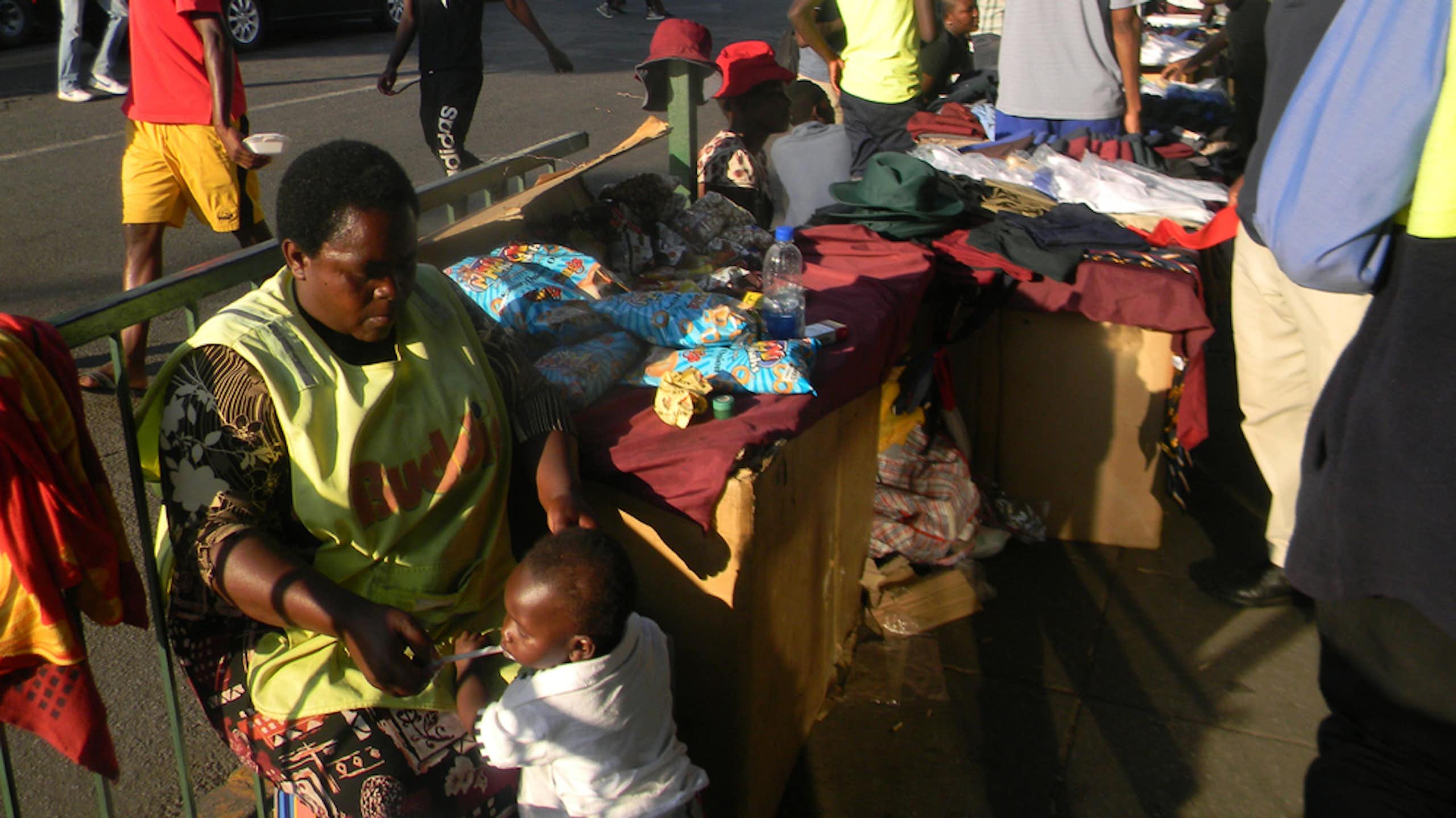Street scene with pedestrians and a woman seated with a display of clothing