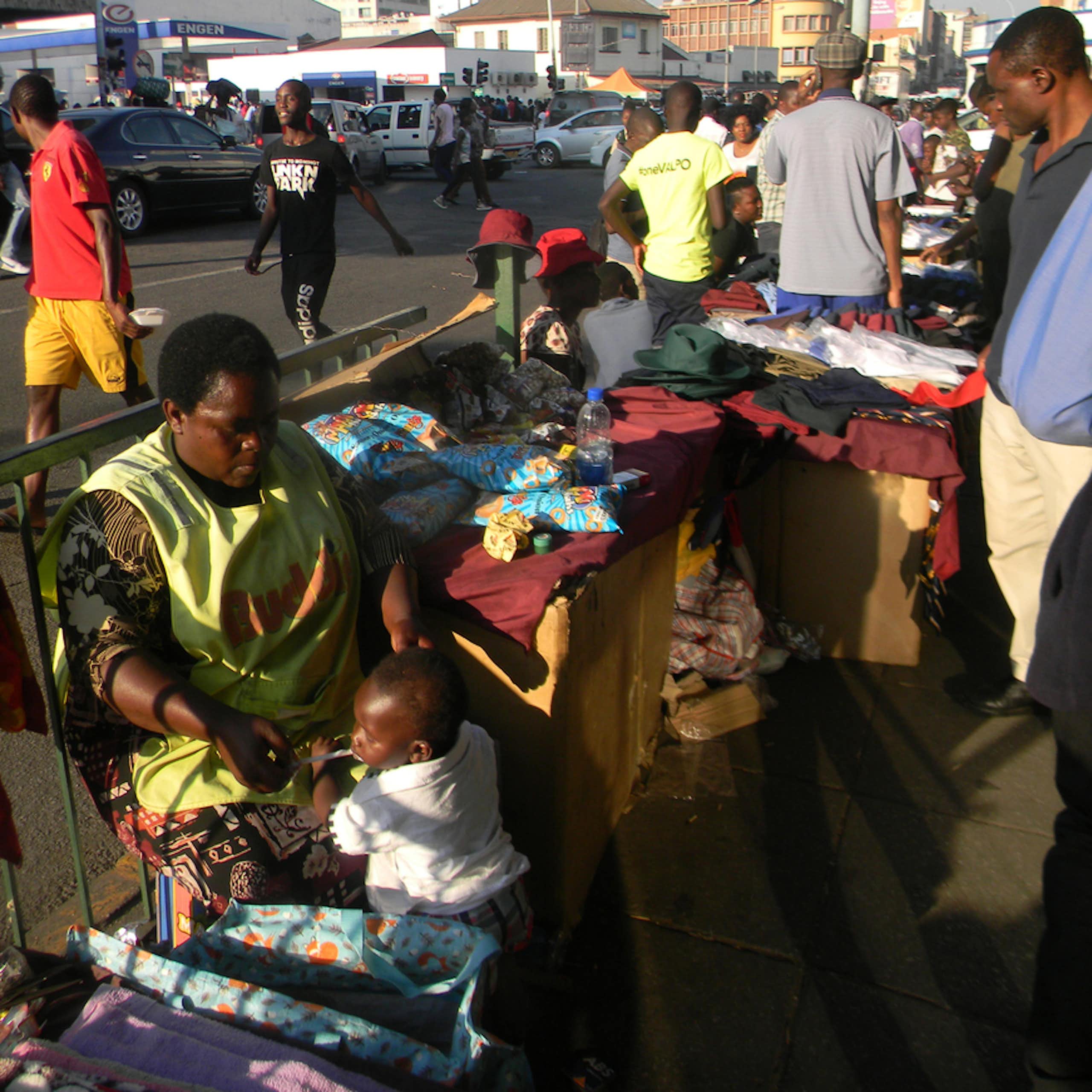 Street scene with pedestrians and a woman seated with a display of clothing