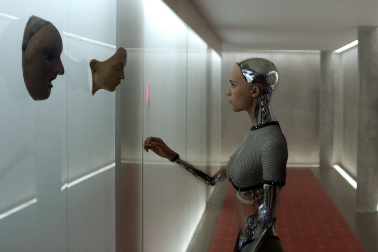A female robot looking at a synthetic face hanging on a wall.
