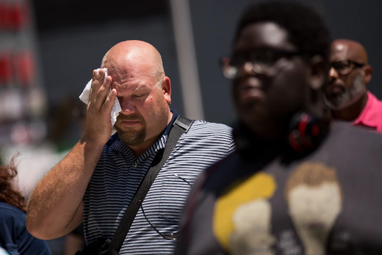 A man walking on a sidewalk wipes his forehead on a hot day.