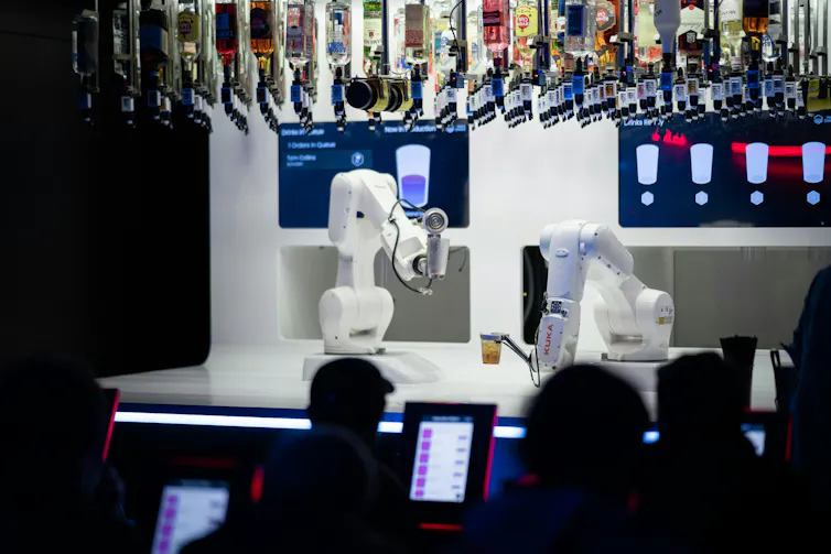 Two bartending robots hold cups while sitting on a bar counter