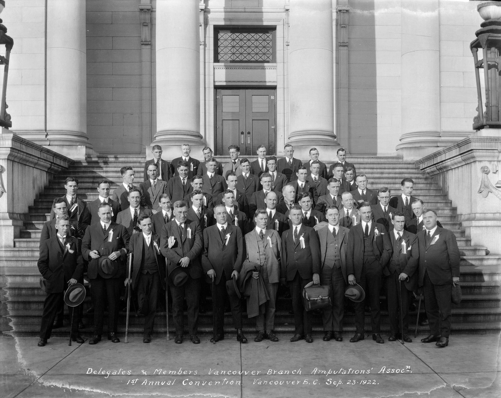 Rows of men standing in suits on steps, many holding one crutch, who have one leg.