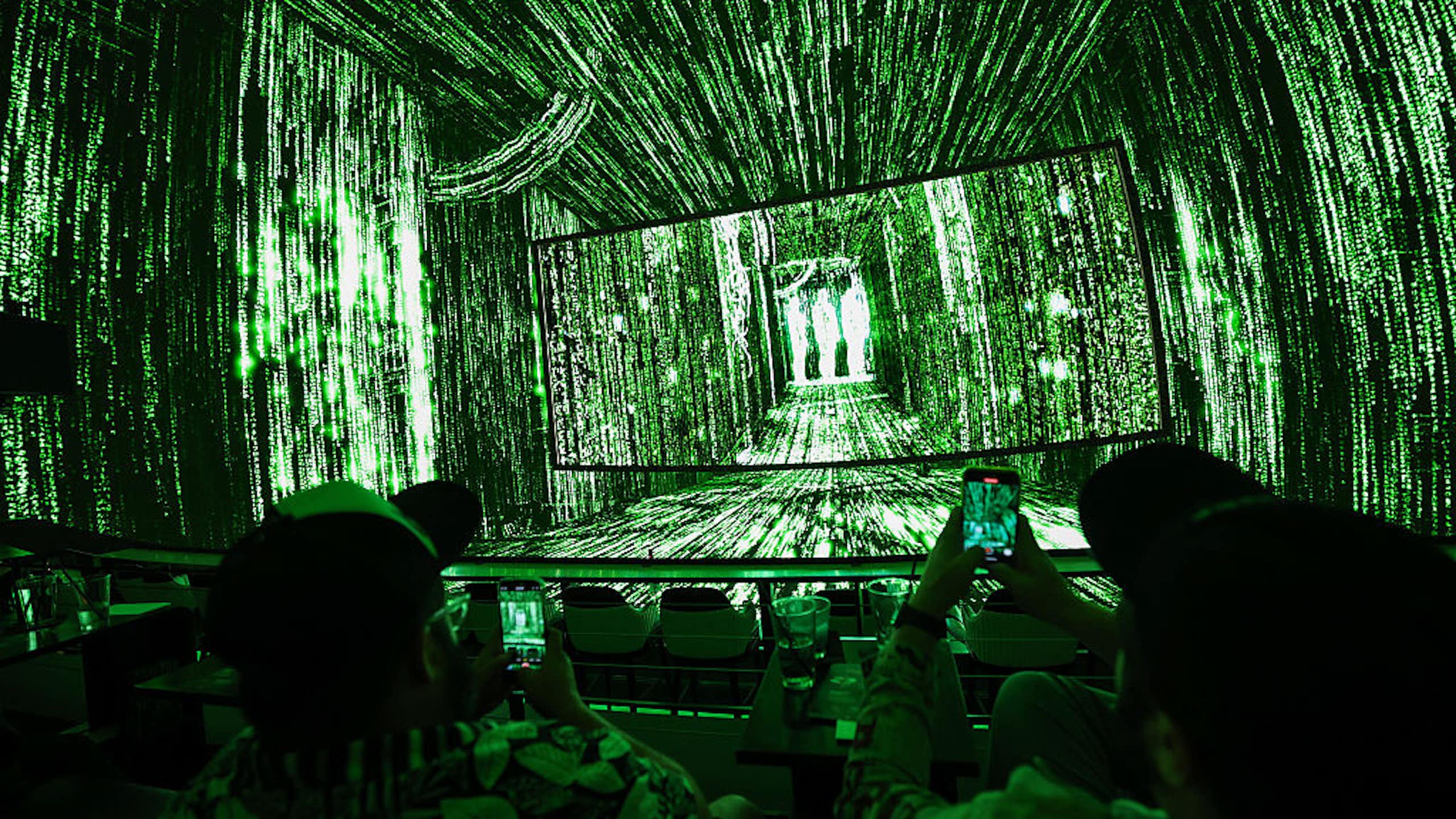 People sit in a darkened room whose walls and ceilings seem to be screens with neon green strings of numbers flashing across them.
