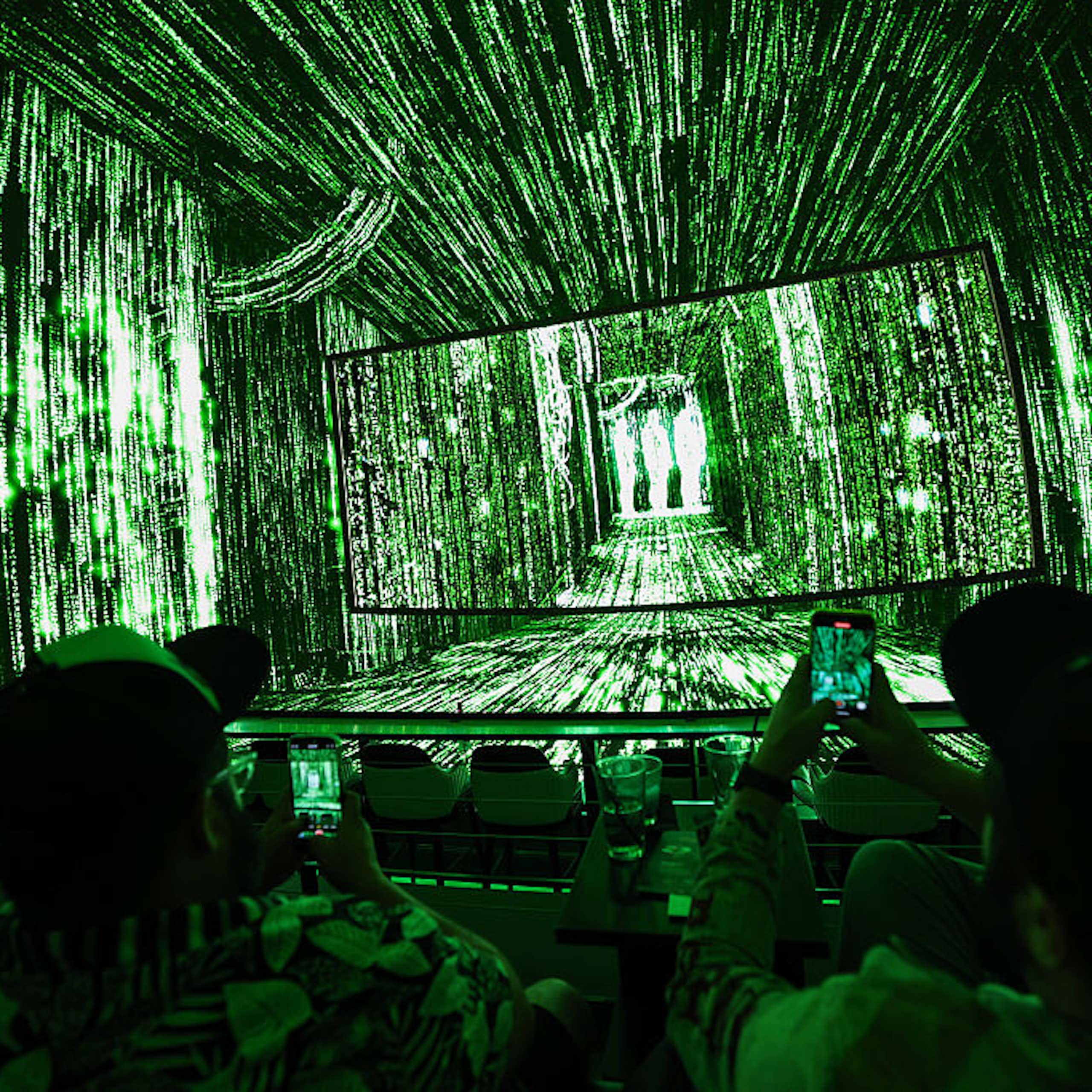People sit in a darkened room whose walls and ceilings seem to be screens with neon green strings of numbers flashing across them.