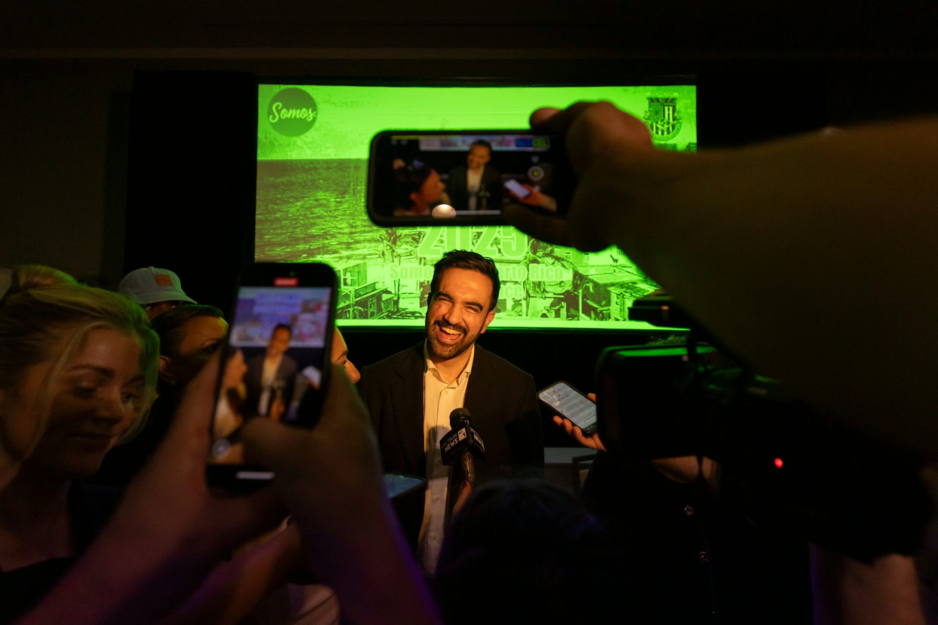A dark-haired man with a beard smiles broadly as people record him on their phones in the foreground.