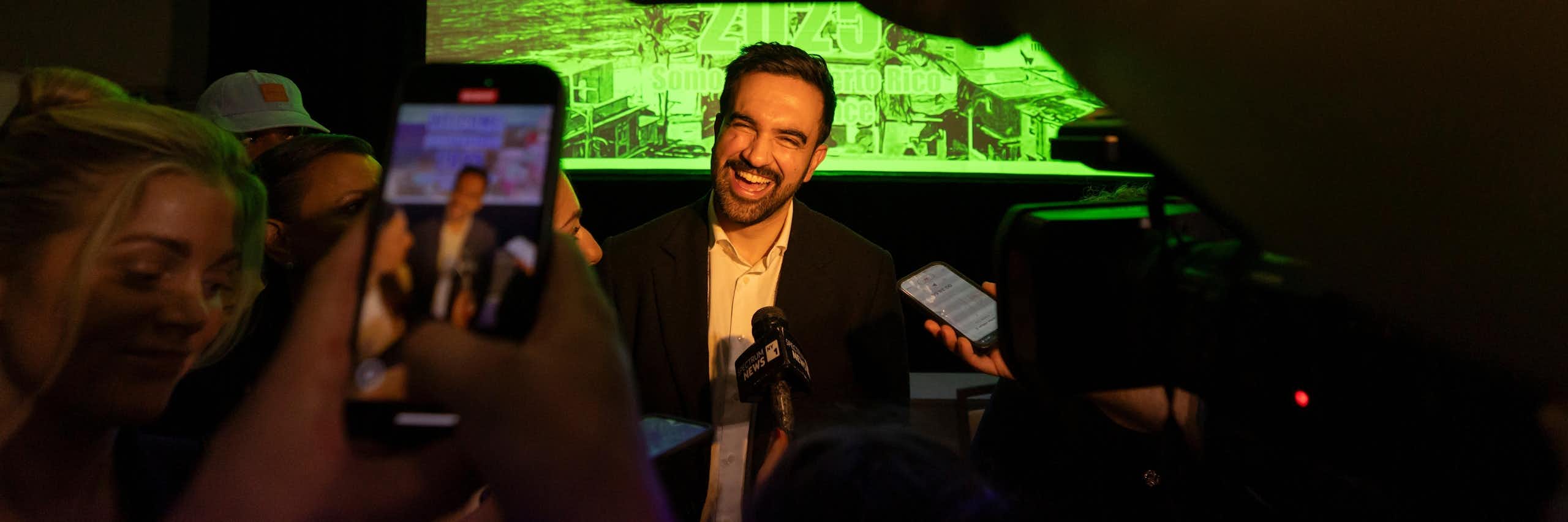 A dark-haired man with a beard smiles broadly as people record him on their phones in the foreground.