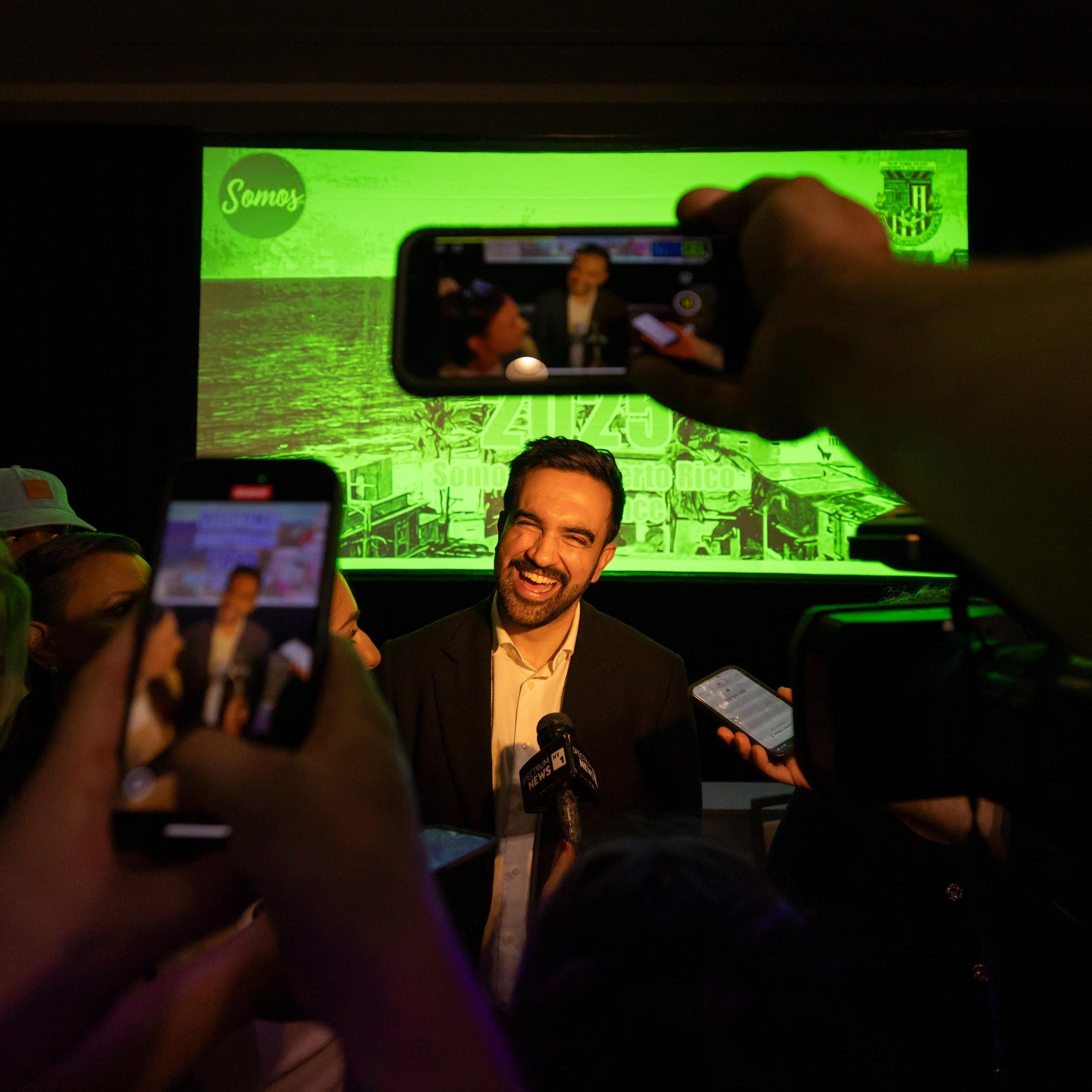 A dark-haired man with a beard smiles broadly as people record him on their phones in the foreground.