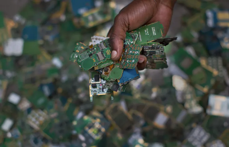 close-up of a hand holding green circuit boards. A pile of other circuit boards is seen in the background.