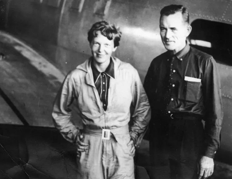 A black and white photo of American pilot Amelia Earhart and her navigator, Captain Fred Noonan, standing in front of a plane in a hangar.
