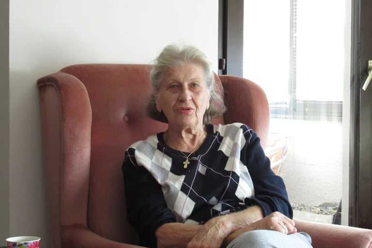An older woman sits in an armchair next to a window