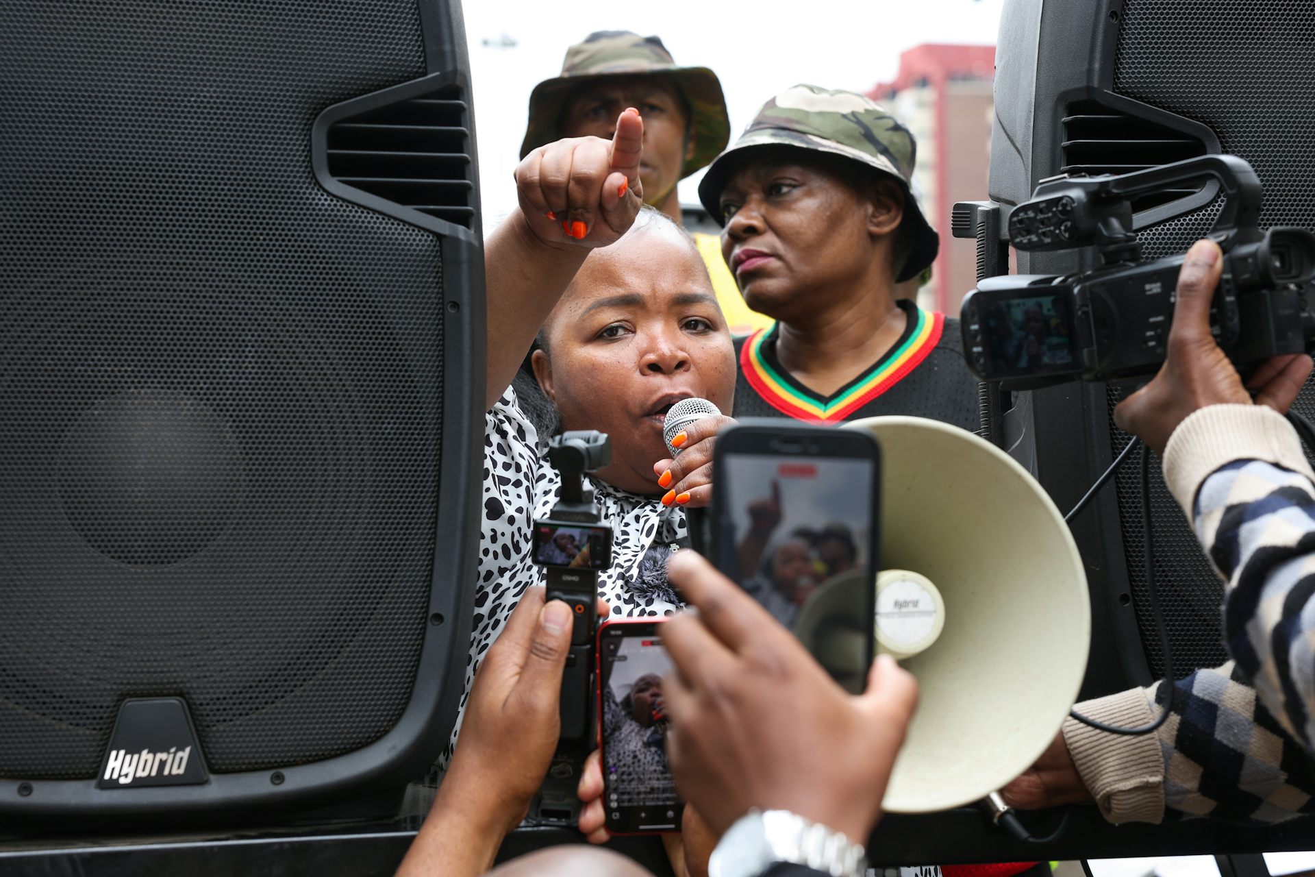 A woman speaks into a microphone while largely obscured by two large speakers on either side of her and a camera phone taking her video
