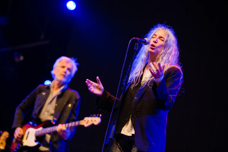 The singer Patti Smith on a black stage, spotlit with a guitarist behind her.