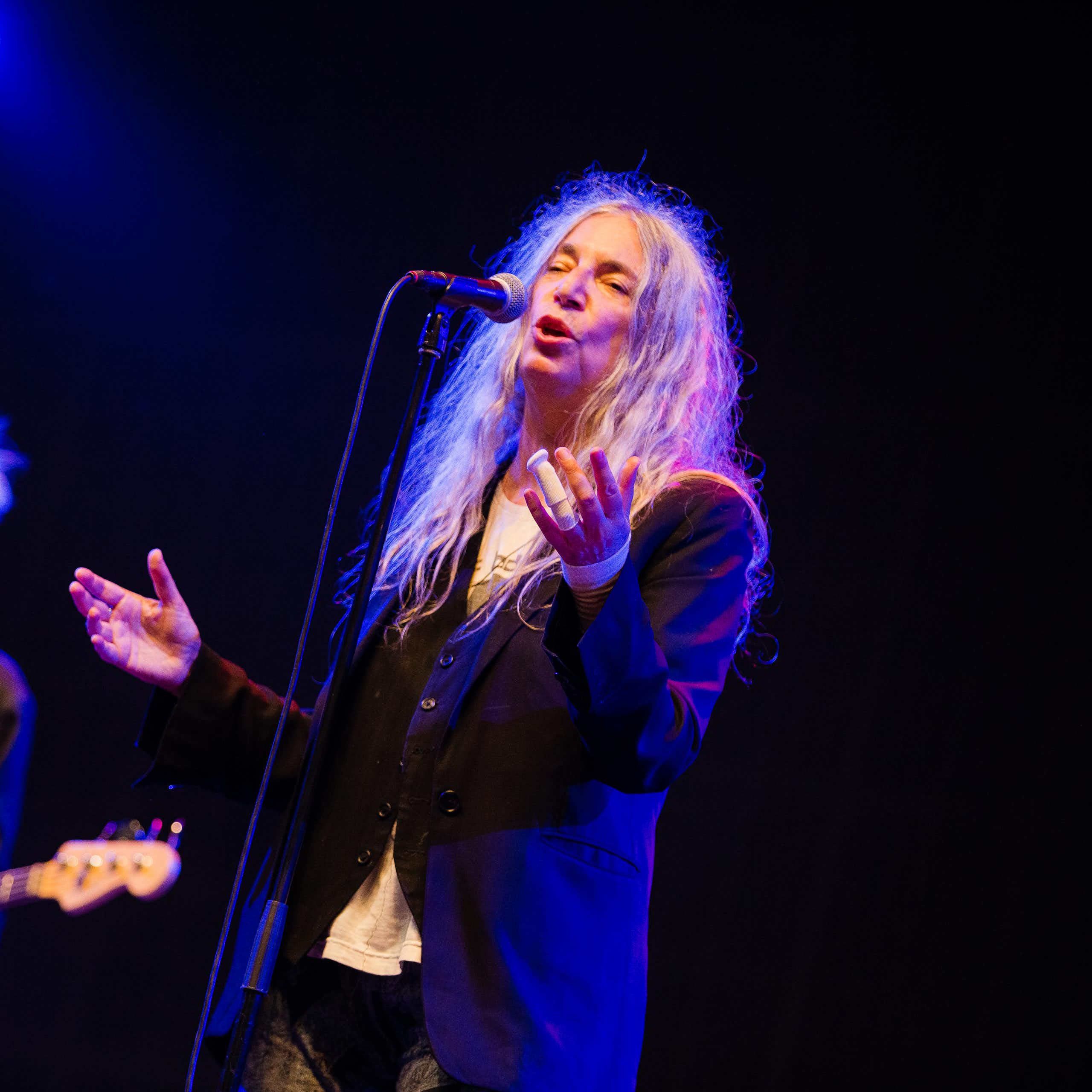 The singer Patti Smith on a black stage, spotlit with a guitarist behind her.