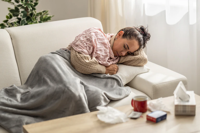 woman resting under a blanket surrounded by tissues, medication and a hot drink