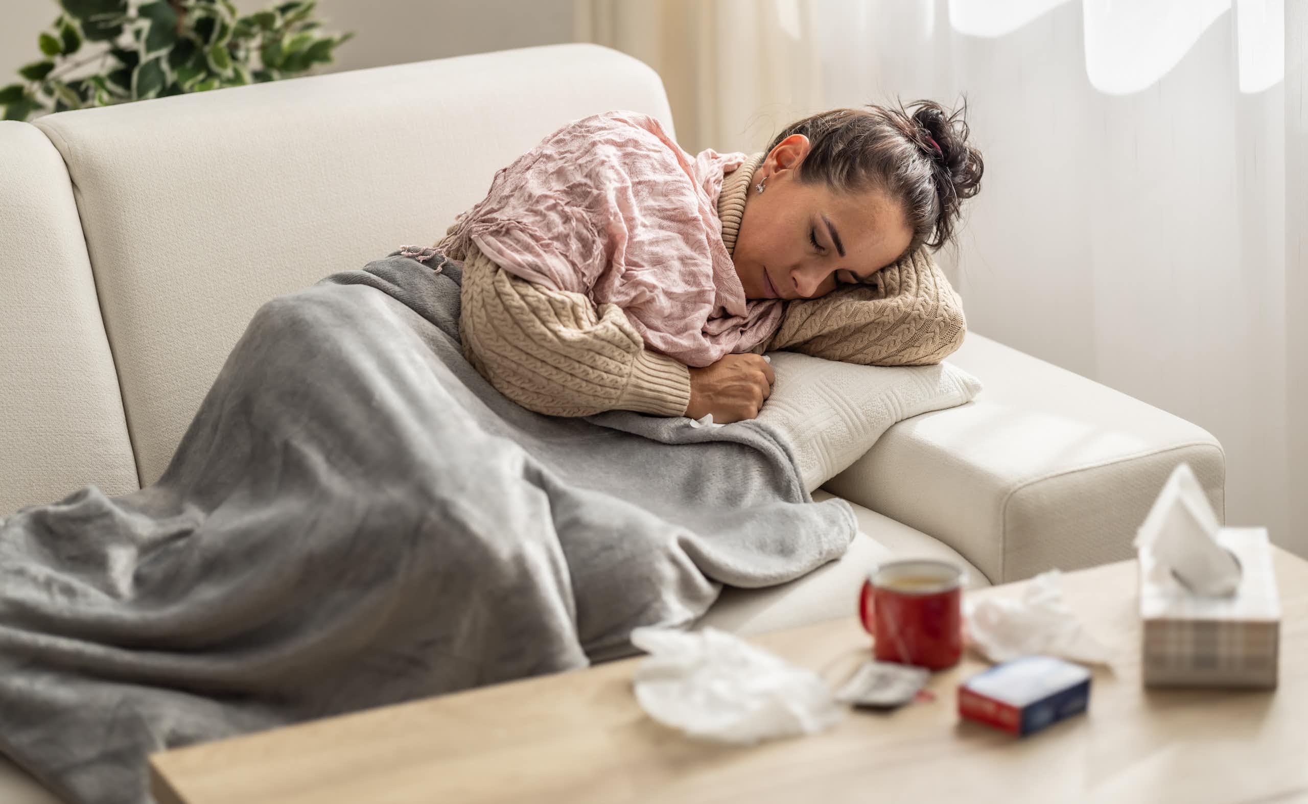 woman resting under a blanket surrounded by tissues, medication and a hot drink