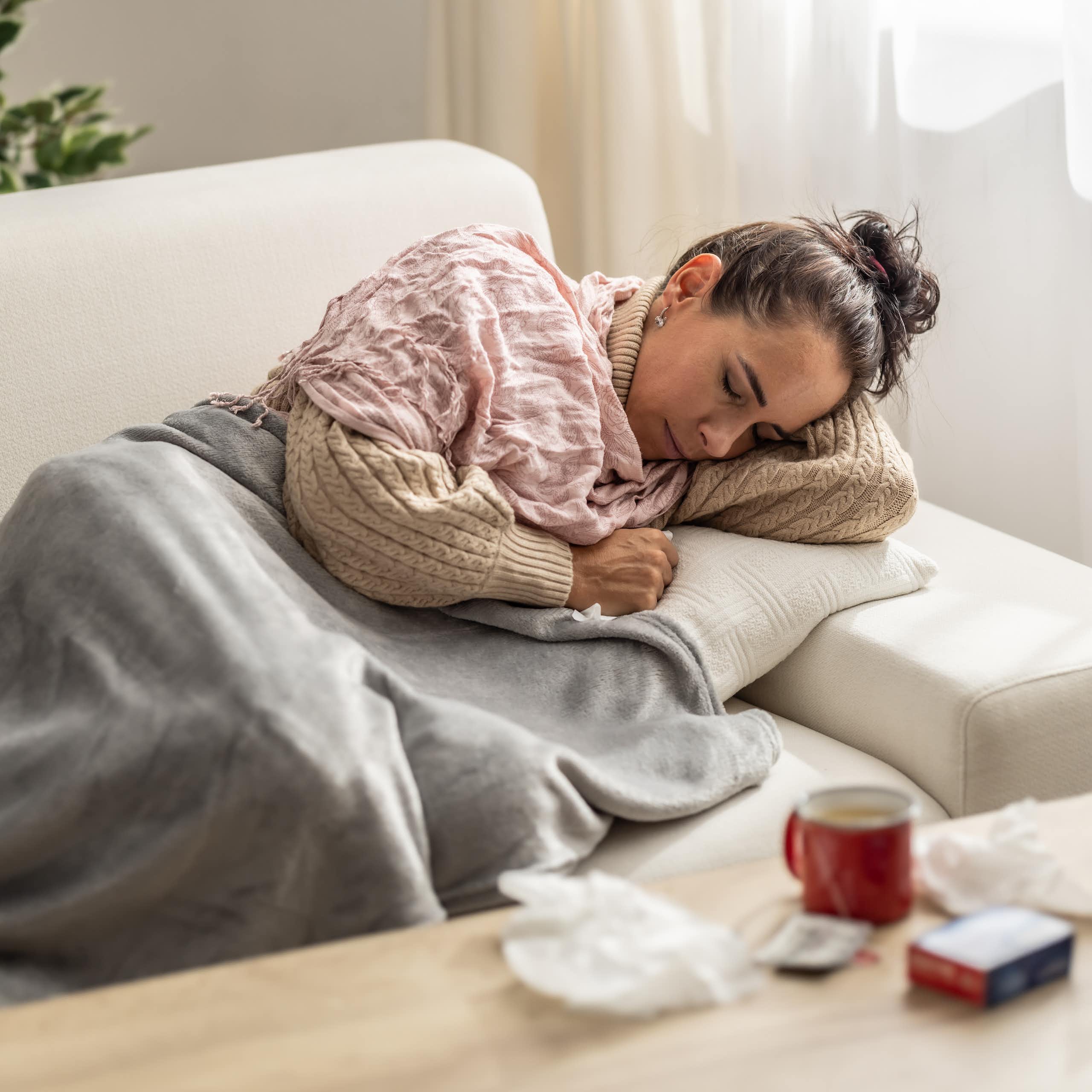 woman resting under a blanket surrounded by tissues, medication and a hot drink