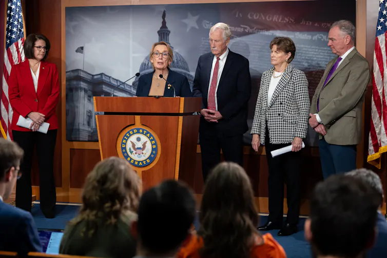 Three women and two men on a stage with American flags flanking them, and one of them speaking at a lectern.