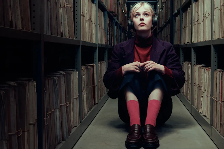 A young blonde woman wearing headphones sits in a narrow hallway surrounded by stacks of shelved files.