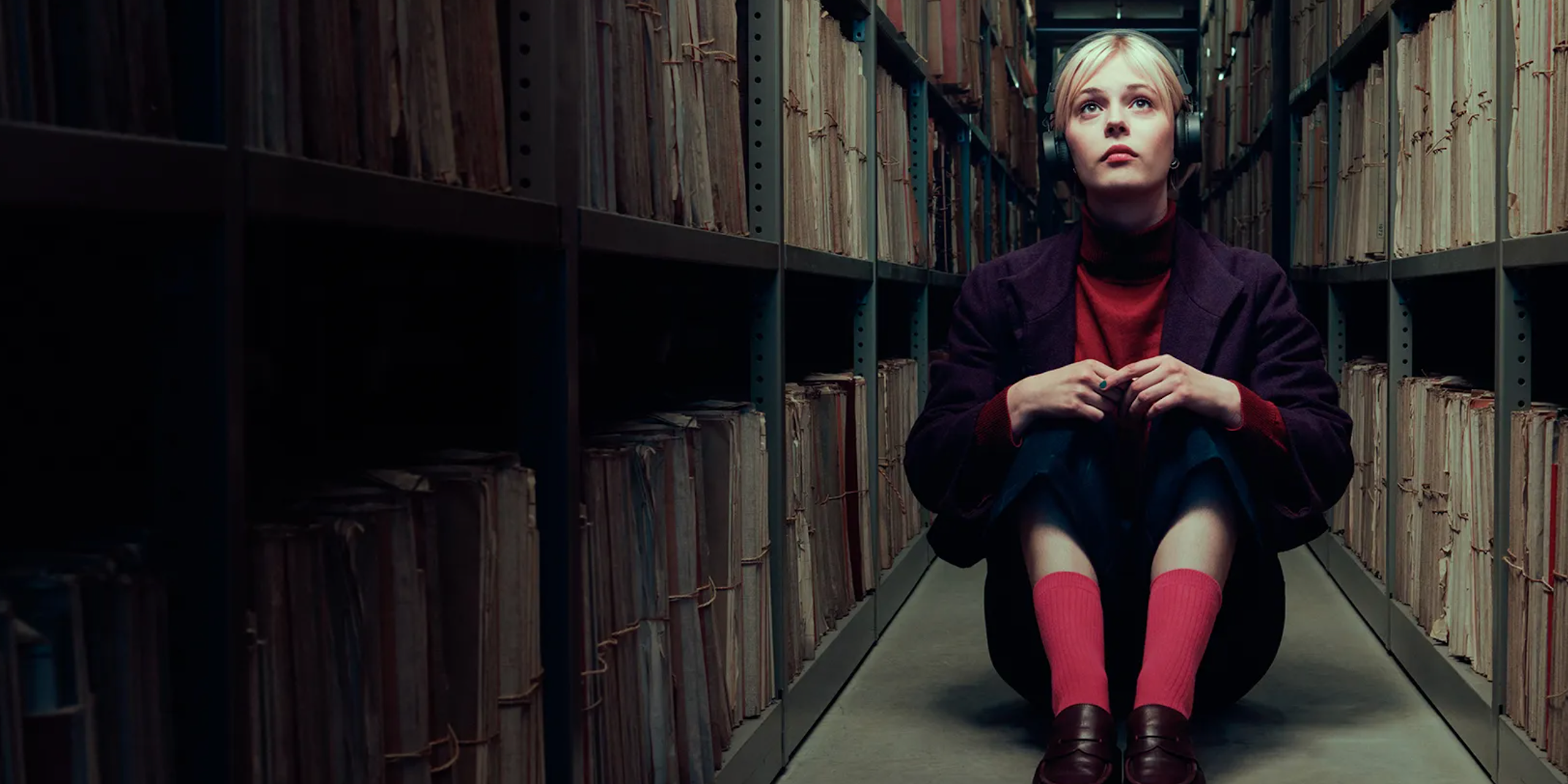 A young blonde woman wearing headphones sits in a narrow hallway surrounded by stacks of shelved files.