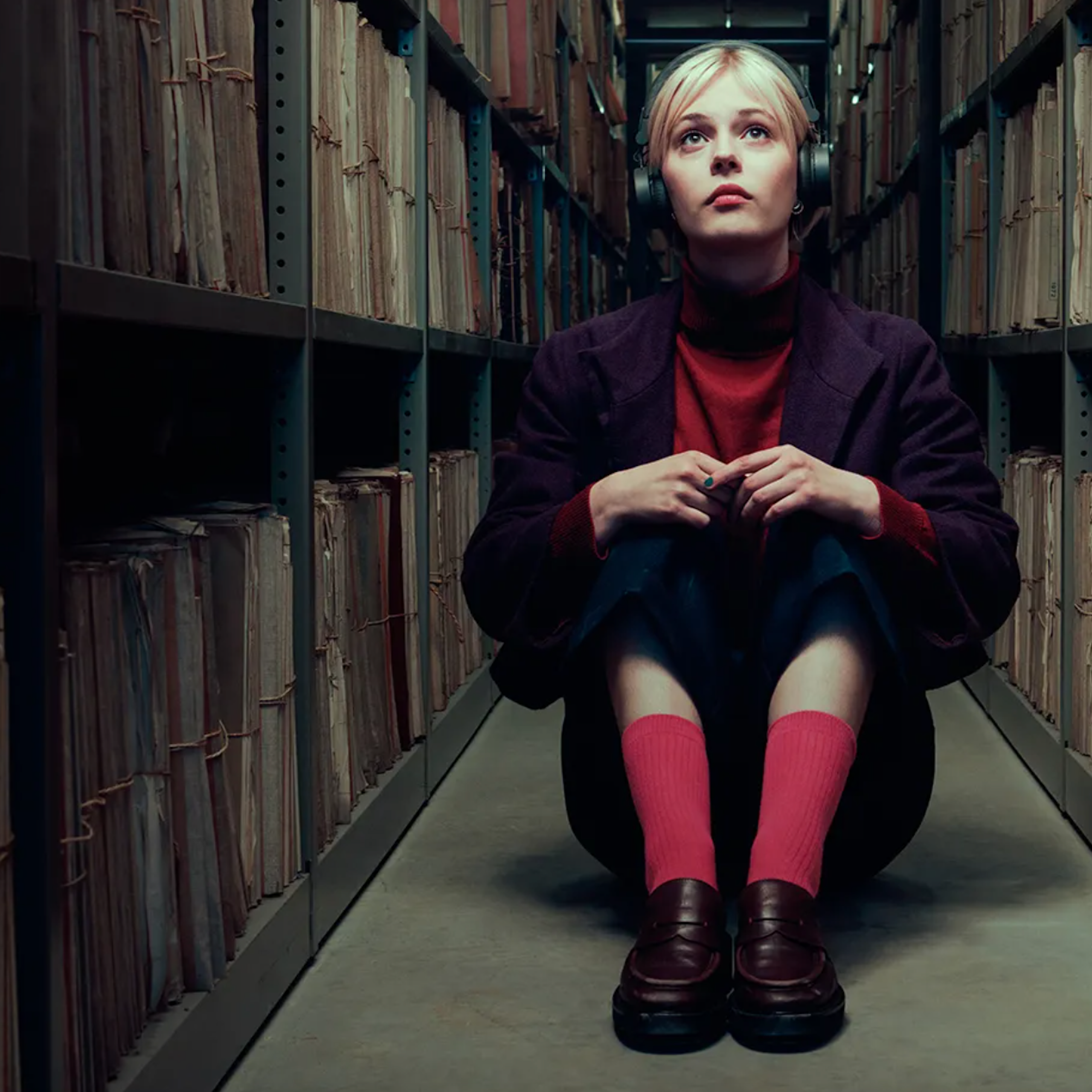 A young blonde woman wearing headphones sits in a narrow hallway surrounded by stacks of shelved files.
