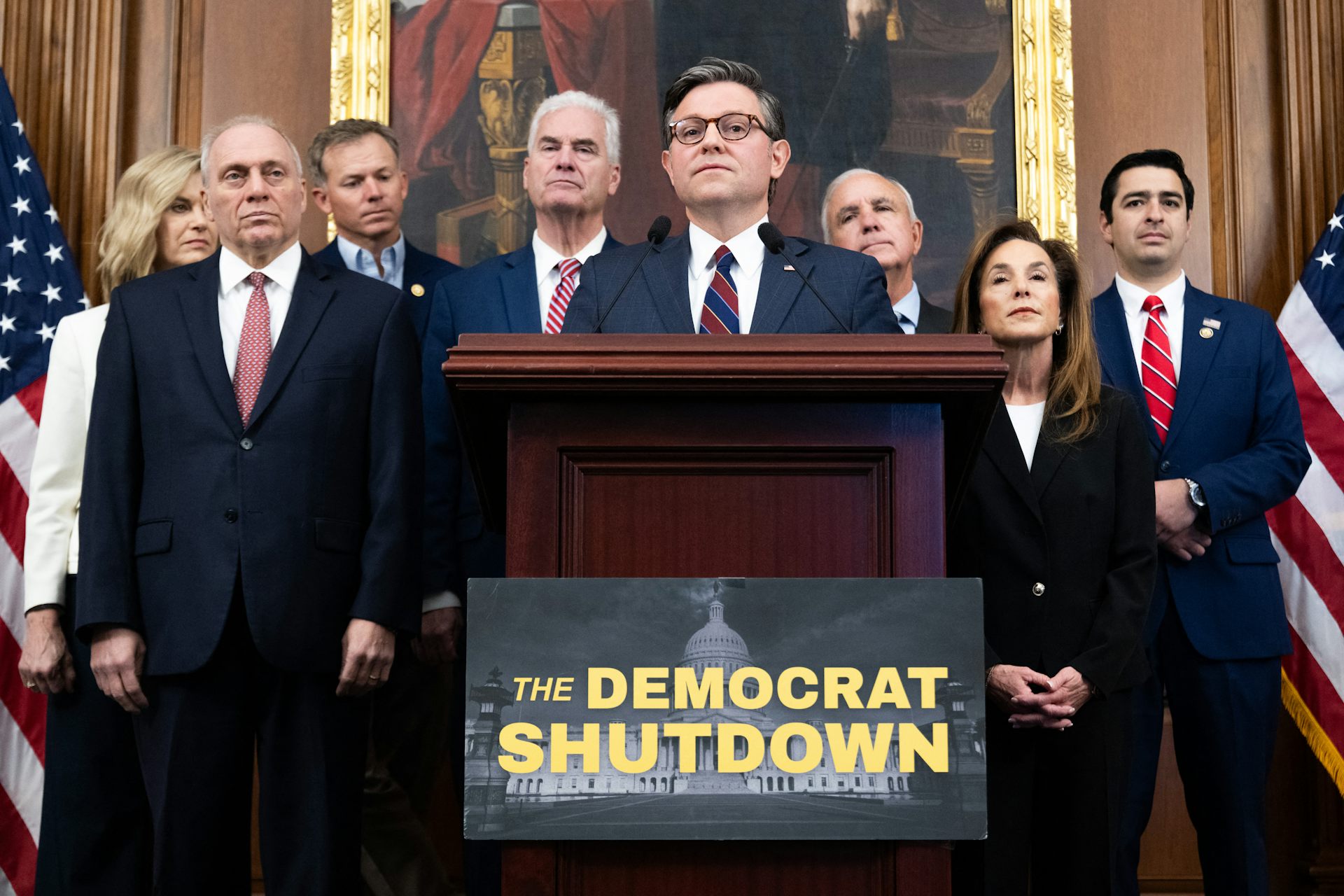 A group of people standing behind a man who's standing at a lectern, behind a sign that says 'The DEMOCRAT SHUTDOWN.'
