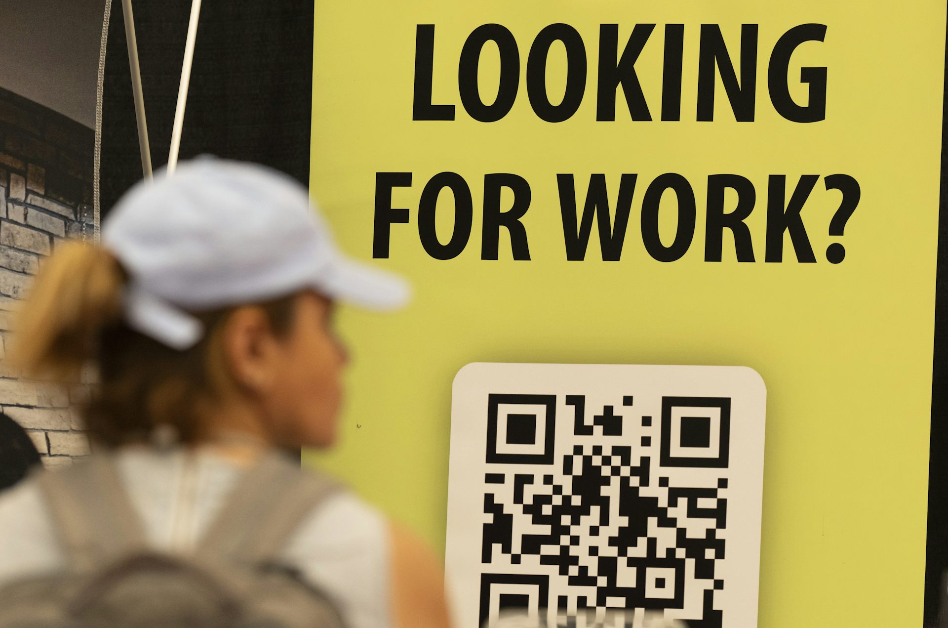 A yellow sign reads 'Looking for Work' with a QR code underneath the words and a woman in a ball cap in the foreground.