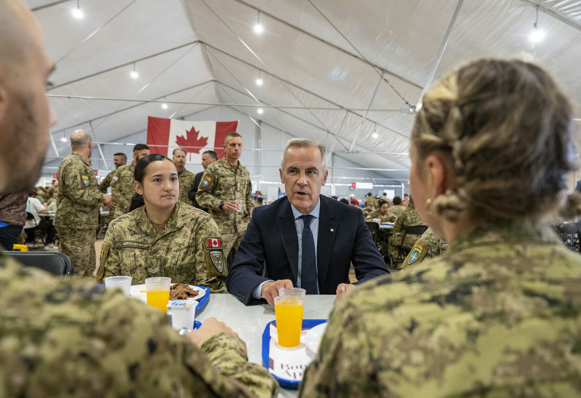 A man with short grey hair sits among soldiers in uniform in a mess hall.