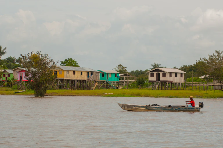 Um barco navega por um rio com casas coloridas na margem ao fundo.