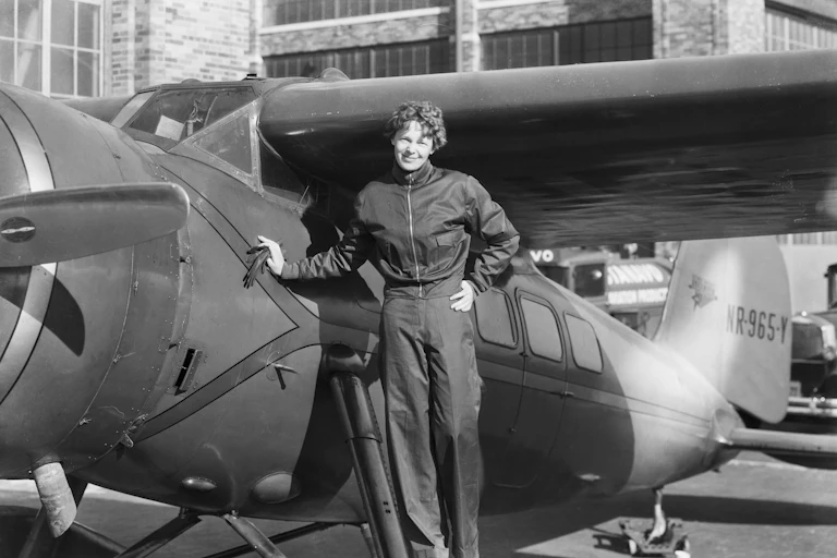 A black and white photo of American pilot Amelia Earhart, smiling, while standing next to an aircraft in her pilot's outfit.