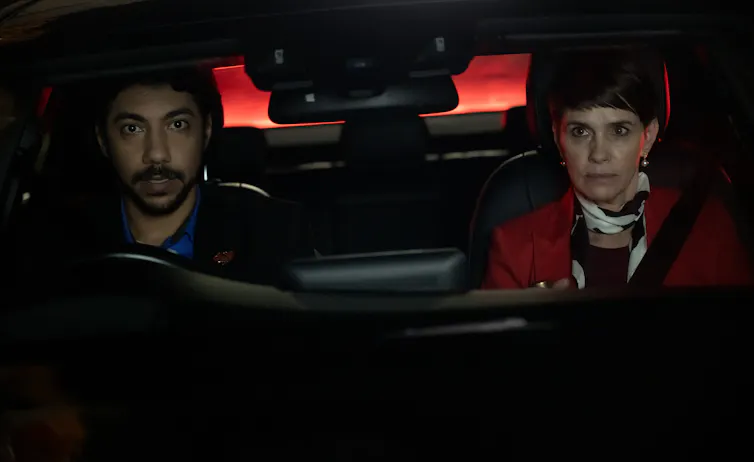A man and woman are in the front seats of a car, looking out. It is night time, and they have serious expressions.