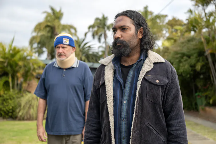 Two men stare at something offscreen while standing in a grassy field. There are palms in the background, and the man on the left has a neck brace.