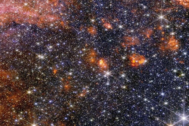 A wide view of a region of space filled with stars and clumps of orange clouds as seen by the James Webb Space Telescope.